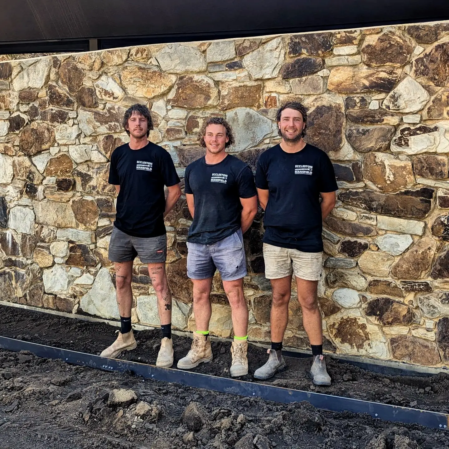 Three Eccleston Mansfield masons in company shirts, standing in front of a newly laid sandstone wall with their boots in the dirt.