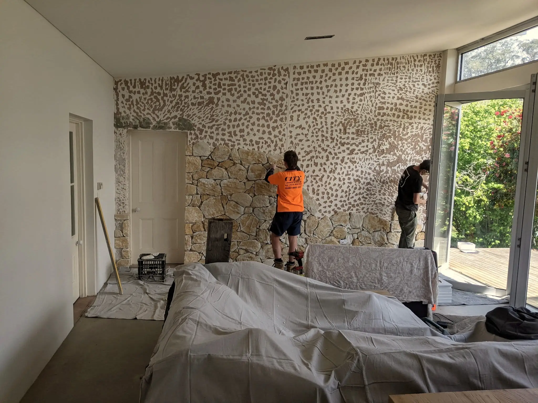 Two masons setting stone on an interior feature wall, floor sheeted and furniture covered.